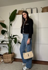Jeune femme en veste noire et jean tient un sac à main beige, debout à l'intérieur près d'une grande plante verte et de placards blancs.