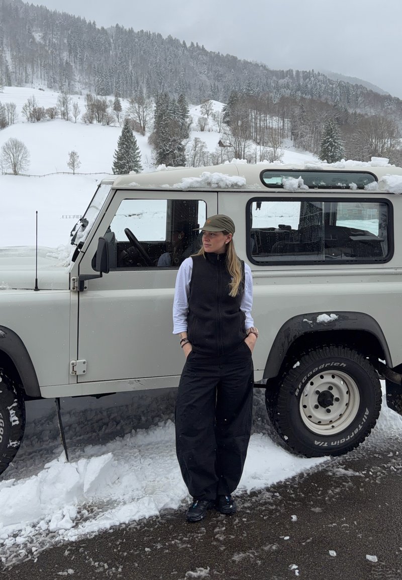 Woman wearing a cap and black vest stands beside a snow-covered white off-road vehicle on a snowy mountain road.