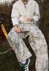 Kremfarget jakke med glidelås kombinert med løstsittende bukser med abstrakt mønster i beige og grønt. Grønne joggesko med hvite striper og synlige tatoveringer på hendene.