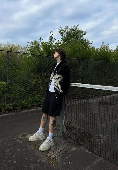 Sudadera negra con cremallera y gráfico blanco, camiseta blanca, pantalones cortos negros, zapatillas blancas gruesas y tatuajes visibles, de pie junto a una valla.