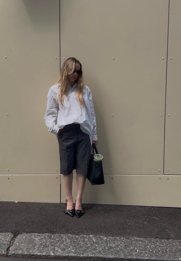 Femme portant une chemise blanche, un short foncé au genou et des talons noirs, debout contre un mur beige tenant un sac noir avec une boisson verte à l'intérieur.