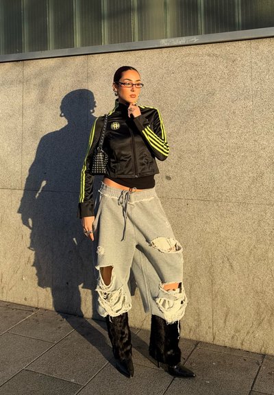 Chaqueta corta negra con rayas neón amarillas, pantalones de chándal grises rasgados y botas de piel negra. Los accesorios incluyen un pequeño bolso con tachuelas y gafas.