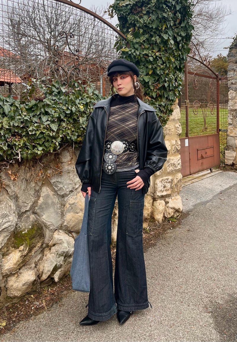 Jeune femme portant un béret noir, une veste noire surdimensionnée, un haut à motifs, un jean évasé, des bottes pointues, tenant un sac fourre-tout en denim près d'un mur en pierre.