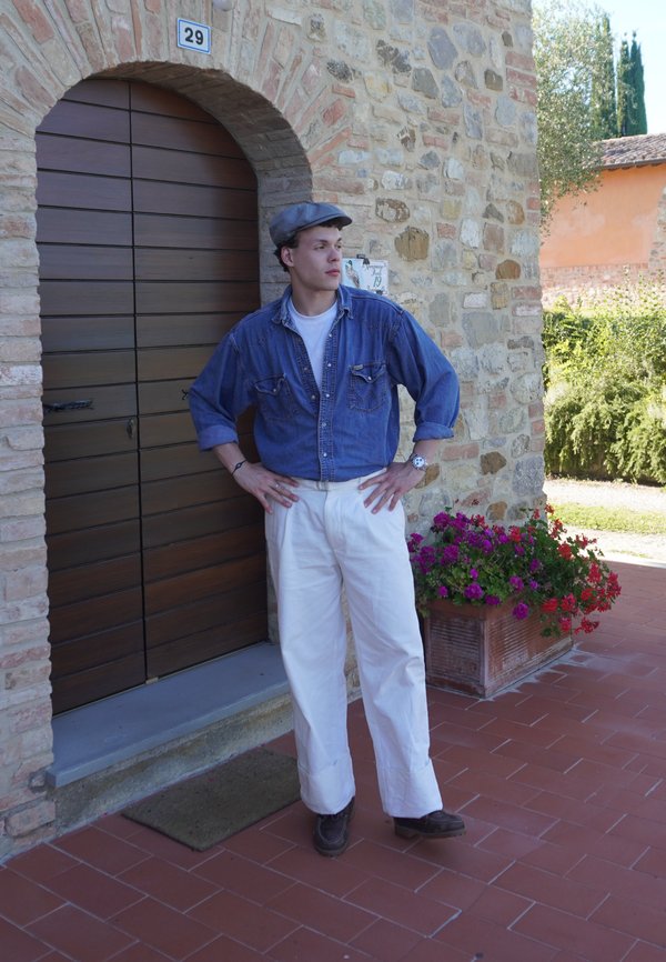 Junger Mann in blauem Jeanshemd, weißen Hosen und Basecap steht mit den Händen in die Hüften vor einem Steingebäude mit Holztür und Blumen.