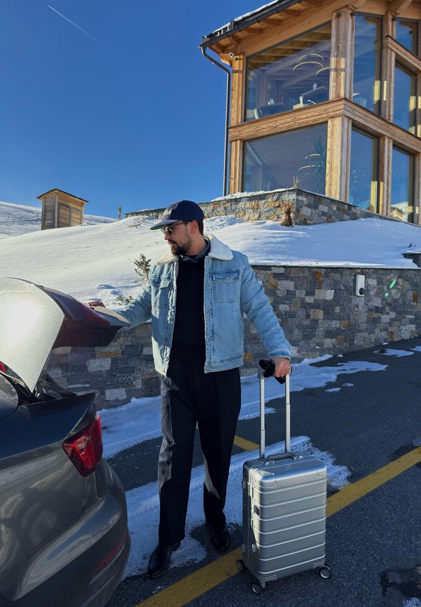 Silberner Hartschalenkoffer mit horizontalen Rillen und ausziehbarem Griff, neben einem schwarzen Auto mit geöffnetem Kofferraum in einer verschneiten Umgebung.