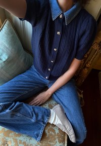 Pull-over à manches courtes en tricot torsadé bleu marine avec col en denim et boutons argentés, assorti à un jean bleu clair et des chaussures beige à enfiler.
