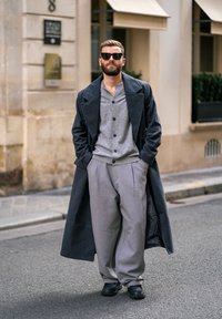 Um casaco cinza escuro sobre um cardigan cinza claro, calças cinza plissadas e sapatos pretos. Acessórios incluem óculos de sol escuros. Ambiente urbano.