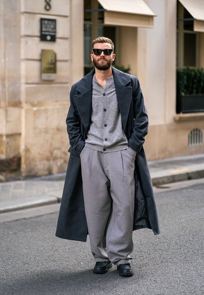 Abrigo gris oscuro sobre un cárdigan gris claro, pantalones gris plisados y zapatos negros. Accesorios con gafas de sol oscuras. Entorno urbano.