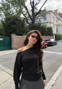 Maglione grigio scuro con scollo a spalle scoperte, orlo e polsini a coste, abbinato a pantaloni grigi plissettati a vita alta. Il modello indossa occhiali da sole e ha capelli ondulati.