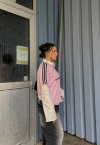 Roza atletska majica s črnimi črtami in znakom, oblečena čez belo s dolgimi rokavi, v kombinaciji s temno sivo ohlapno hlačami.