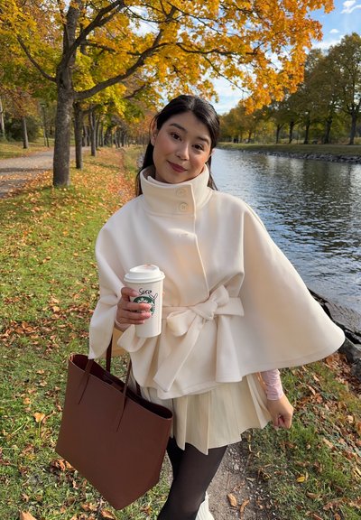 Capa blanca ajustada con gran cuello y cintura atada, llevada sobre una falda clara; sosteniendo una taza de café y un bolso marrón, con un paisaje pintoresco de fondo.