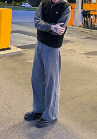 Pantaloni largi gri și o vestă din fleece neagră, asortate cu pantofi cu platformă negri. Fundal din beton cu pompe de combustibil și un obiect portocaliu.