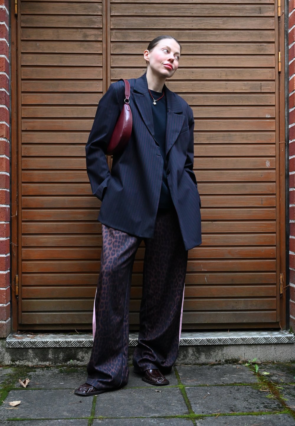 Blazer noir à rayures surdimensionnées sur un haut noir, associé à un pantalon imprimé léopard marron. Accessoirisé avec un sac bordeaux et une paire de chaussures sombres.
