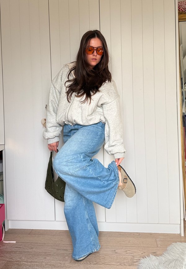 Vrouw met oranje zonnenbril, grijze oversized trui, blauwe jeans met wijde pijpen, houdt een handtas vast, past haar beige sandaal binnen aan.