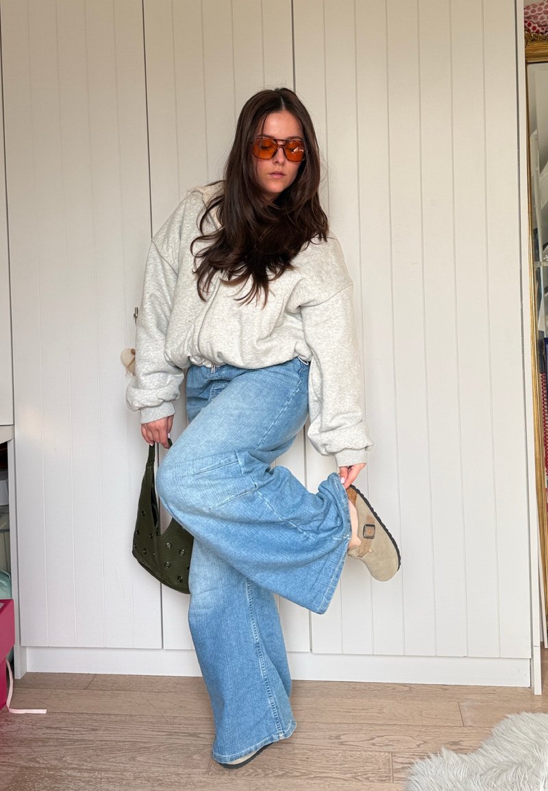 Woman wearing orange sunglasses, gray oversized sweater, blue wide-leg jeans, holding a handbag, adjusting her beige sandal indoors.