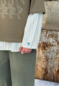 T-shirt oversize vert olive avec un motif d'animal brodé blanc, portée sur une chemise à rayures bleues et blanches avec boutons. Poignet ajustable avec détail en forme de cœur.