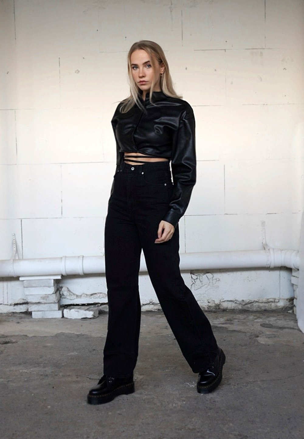 Chaqueta corta de cuero sintético negra con un cuello alto, combinada con vaqueros negros de cintura alta y zapatos negros gruesos. Fondo liso.