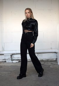 Chaqueta corta de cuero sintético negra con un cuello alto, combinada con vaqueros negros de cintura alta y zapatos negros gruesos. Fondo liso.