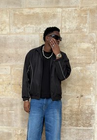 Czarna kurtka zapinana na zamek z białymi lamówkami, czarna koszula i luźne niebieskie dżinsy. Szczególne akcesoria to okulary przeciwsłoneczne, naszyjnik i pierścionki.