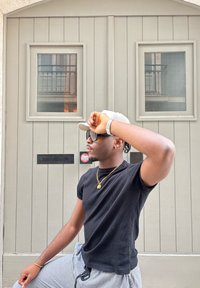 Tricou negru cu dungi, pălărie gri deschis și ochelari de soare. Ușă din lemn gri cu două feronerie dreptunghiulare în spate. Pantaloni ușori în carouri.