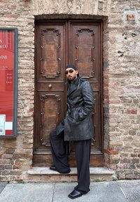 Homme portant un manteau en cuir noir, des lunettes de soleil et un foulard, appuyé contre une porte en bois ornée avec un mur en briques en arrière-plan.