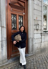 Granatowa marynarka, kremowe spodnie o szerokich nogawkach, trzymając beżową torbę. W tle znajduje się drewniane drzwi z misterną żeliwną kratą i kamienną ścianą.