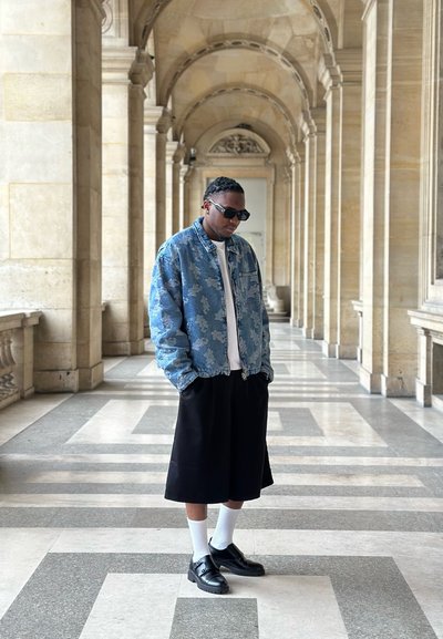 Hombre con gafas de sol, chaqueta vaquera estampada, camisa blanca, pantalones cortos negros anchos, calcetines blancos y zapatos negros, de pie en un corredor de piedra con columnas.