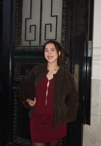 Braune Wildlederjacke über einem burgunderroten Kleid mit einem Schlitzdetail, kombiniert mit transparenten Strumpfhosen. Im Hintergrund ist eine kunstvoll verzierte schwarze Tür zu sehen.