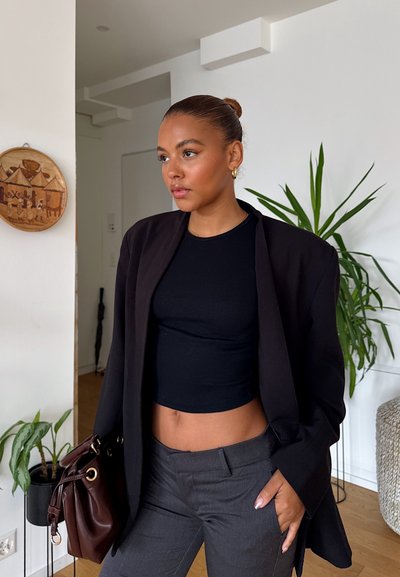 Blazer negro oversize sobre un top corto negro ajustado de canalé, pantalones grises de corte entallado. Bolso marrón con herrajes dorados. Planta verde en el fondo.