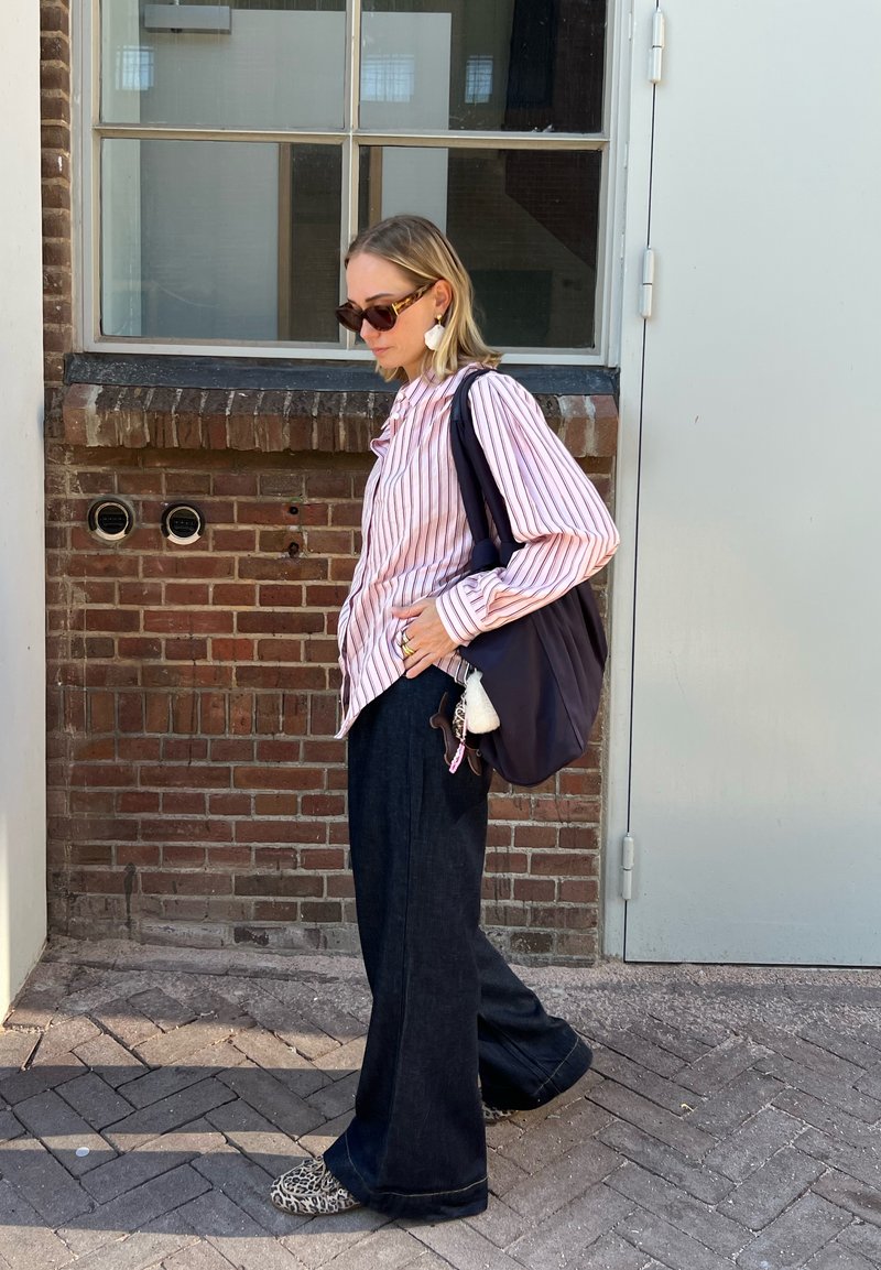 Camisa listrada em rosa e branco com botões, jeans largo escuro, bolsa preta, sapatos com estampado de animal e óculos de sol oversized. Fundo em tijolo.