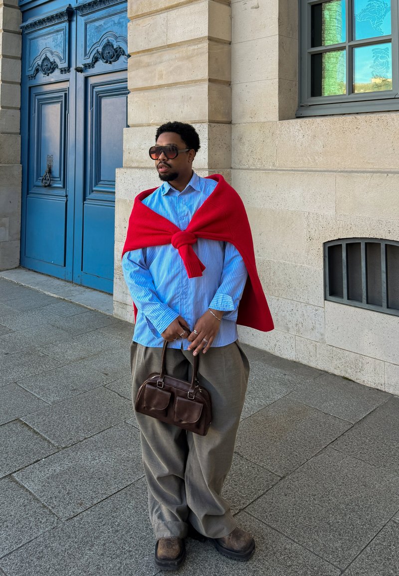 Homme portant des lunettes de soleil, un pull rouge sur une chemise bleue, tenant un sac à main marron, debout sur le trottoir près d’un mur en pierre beige et d’une porte bleue.