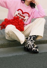 Pull roz cu textul "DIN VIITOR", pantaloni bej cu picioare largi și botine cu model de vacă alb-negru. Ține o geantă texturată în formă de floare roșie.