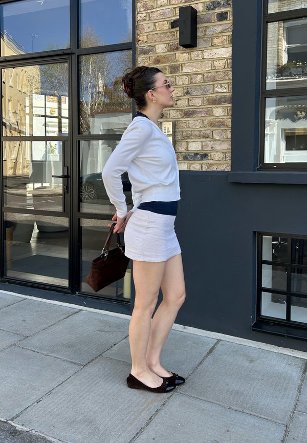 Žena u bijelom kardiganu i suknji stoji na pločniku držeći tamnu torbu iza leđa ispred ciglene zgrade.