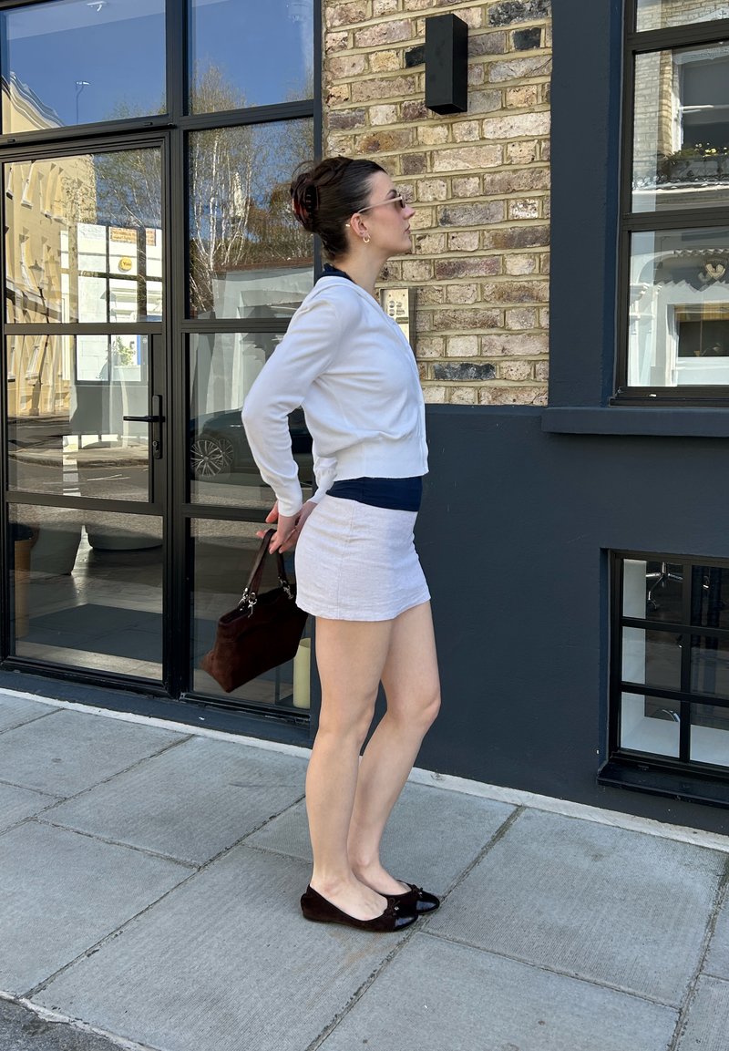 Donna con cardigan bianco e gonna, in piedi sul marciapiede, che tiene una borsa scura dietro la schiena davanti a un edificio in mattoni.