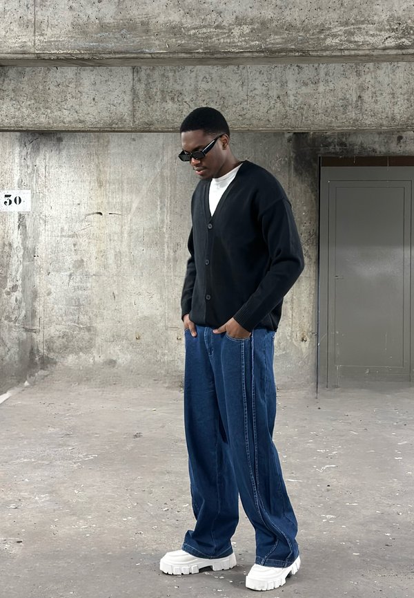 Schwarze Strickjacke mit Knopfverschluss, weißes T-Shirt darunter, lockere blaue Jeans mit seitlichen Streifen und klobige weiße Sneakers.