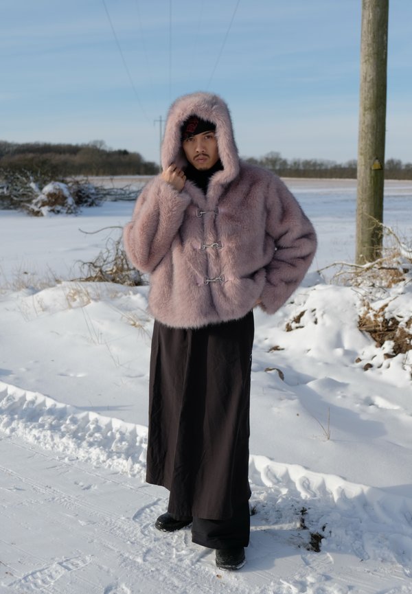 Persoană purtând o haină roz cu glugă din blană și pantaloni negri cu picioare largi, stând în aer liber pe un drum acoperit de zăpadă, cu copaci și linii electrice în fundal.