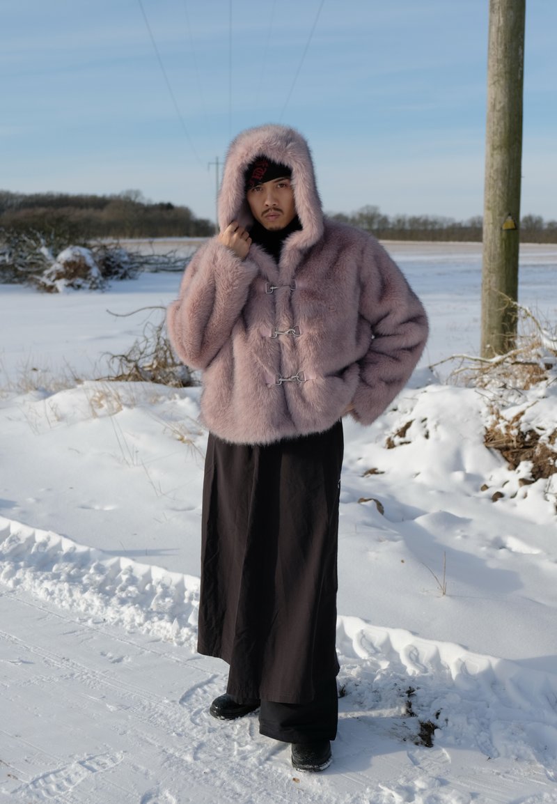 Person som bär en rosa pälsfodrad huva och svarta byxor med vida ben, stående utomhus på en snötäckt väg med träd och kraftledningar i bakgrunden.