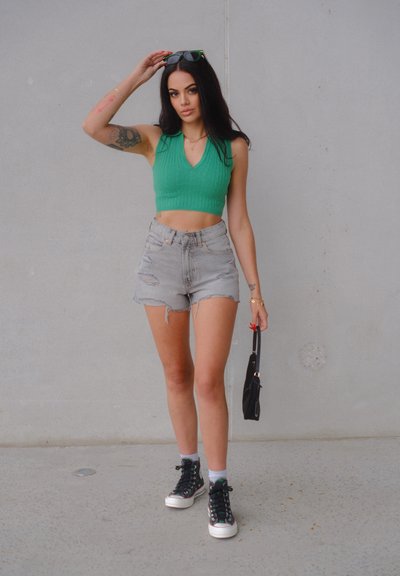 Top corto verde de canalé, pantalones cortos de mezclilla rasgados de tiro alto, zapatillas altas negras con acentos de color y un pequeño bolso negro.