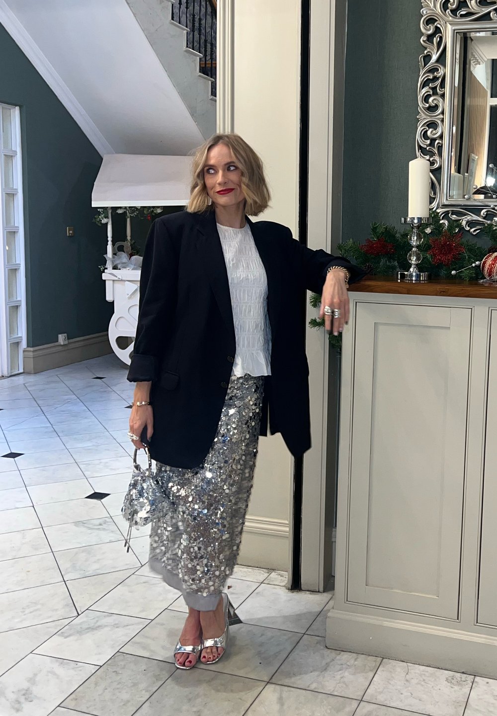 Blazer noir oversize, haut blanc texturé, jupe crayon argentée à paillettes, talons argentés, tenant un petit sac à main, debout dans un espace intérieur bien éclairé.