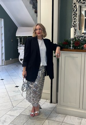 Blazer negro de gran tamaño, top blanco texturizado, falda lápiz plateada con lentejuelas, tacones plateados, sosteniendo un bolso pequeño, posando en un espacio interior bien iluminado.