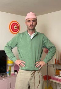 Svetlozelená mikina na zips s dlhými rukávmi, nosená cez biele tričko; doplnená ružovou pletenou čiapkou. V pozadí farebná výzdoba.