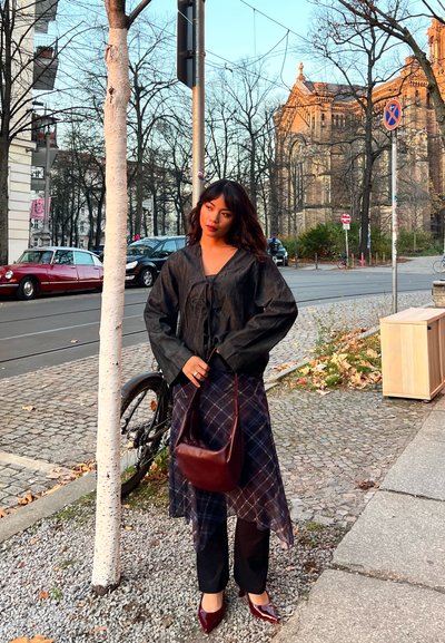 Blusa de lino negra con un lazo frontal, combinada con una falda transparente de cuadros oscuros. Se acompaña de pantalones oscuros y zapatos de punta en color granate. Bolso marrón.