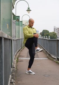 Man i träningskläder som sträcker ut sitt ben på en gångbro med metallegaller och gatlyktor i en urban miljö.