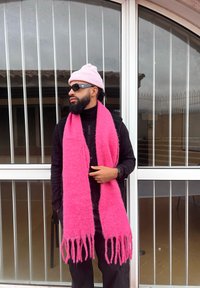 Ροζ μάλλινο κασκόλ με φούντες φορεμένο πάνω από μαύρο πουλόβερ με ψηλό λαιμό. Ένα επιπλέον ροζ σκουφάκι και γυαλιά ηλίου συμπληρώνουν την σκούρα εμφάνιση. Εσωτερικός χώρος.