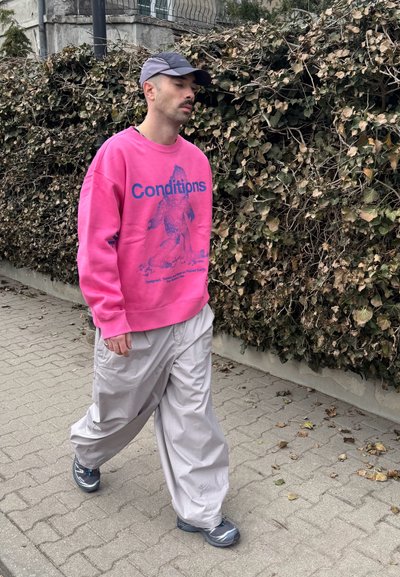 Hombre caminando por la acera con sudadera rosa con el texto "Conditions", pantalones sueltos de color gris claro, zapatillas grises y gorra azul cerca de un seto seco.