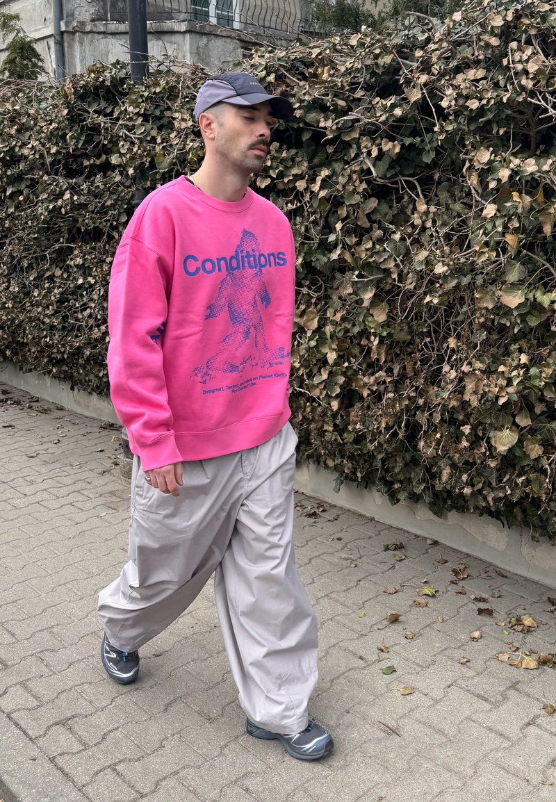 Homme marchant sur le trottoir portant un sweat-shirt rose avec l'inscription « Conditions », un pantalon ample gris clair, des baskets grises et une casquette bleue près d'une haie sèche.