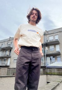 Κρεμ μπλουζάκι από βαμβάκι με το λογότυπο "Carhartt" σε μπλε χρώμα, συνδυασμένο με μαύρο παντελόνι. Ελαφριά, άνετη εφαρμογή; εξωτερικό περιβάλλον.