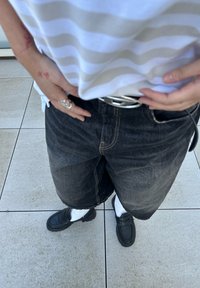 Svarta denimshorts med ett silverbälte, kombinerat med svarta skor och vita strumpor. Skjortan är vit med tunna horisontella ränder.