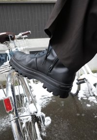 Botte en cuir noir à semelle épaisse posée sur le porte-bagages arrière d'un vélo argenté, sur un trottoir enneigé.