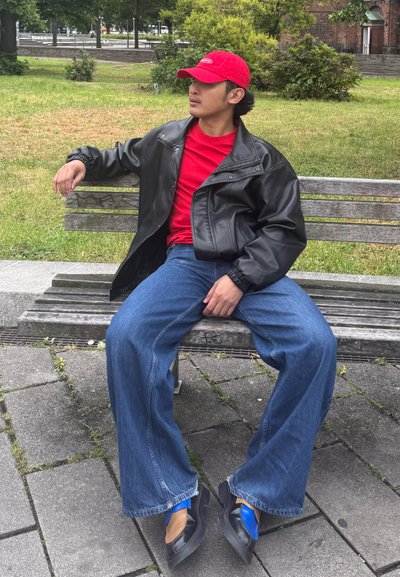 Chaqueta de cuero negra, camisa roja, jeans anchos azules y zapatos negros con acentos azules. Sentado en un banco de madera en una zona de hierba.